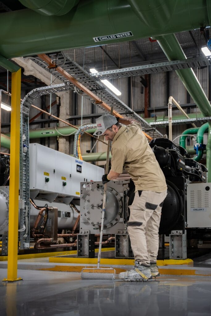 Rolling a grey epoxy coat beside a chiller unit in a commercial plant room